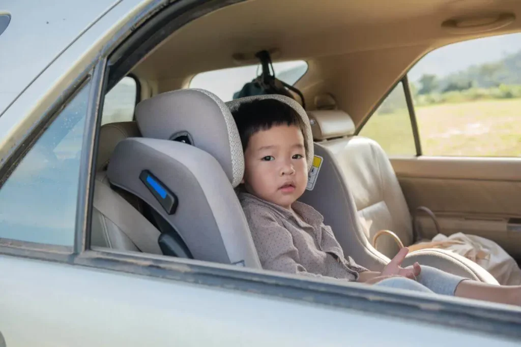 A young child seated securely in a booster car seat inside a vehicle, looking out of the window.