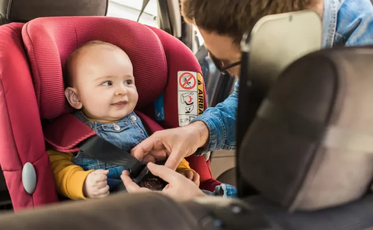 A smiling baby in a red convertible car seat while a father secures the harness inside a car.